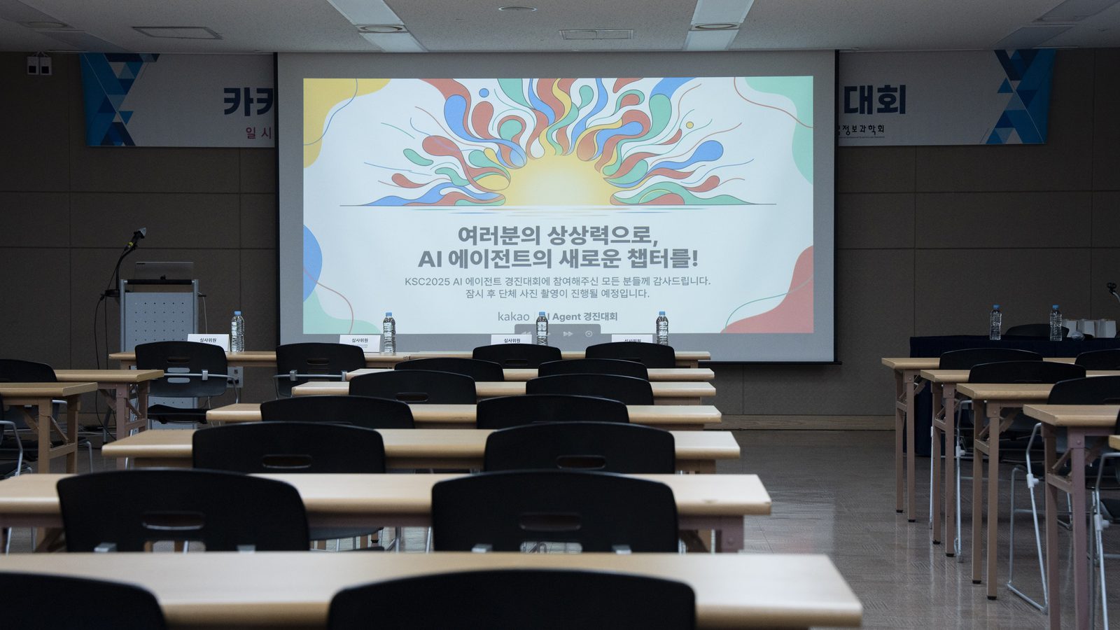 The finals venue of the AI agent competition. Past rows of empty desks, a large screen shows the event's slogan in Korean